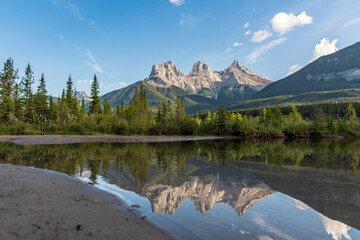 Reflective Three Sisters mountains in Canmore near Banff National Park during summer time with blue sky, lush, green nature, forest woods.