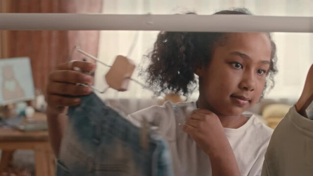 Chest up of cool African American preteen girl looking through clothes on rack while choosing daily outfit in cozy kids bedroom