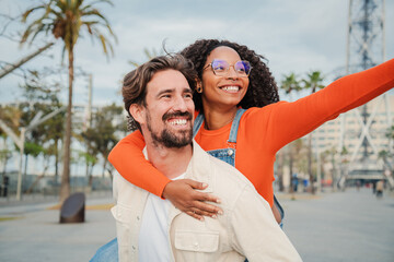 Multiracial couple having fun together on a romantic date. Smiling man giving a piggyback ride to...