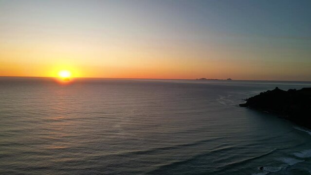 Sunrise over silhouetted islands in the pacific ocean, off the coast of Coromandel, New Zealand. Aerial view, pulling backwards to reveal waves and coastline. 