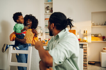 A playful interracial mother and son are playing in a kitchen at home while the father is watching them and looking over the shoulder.