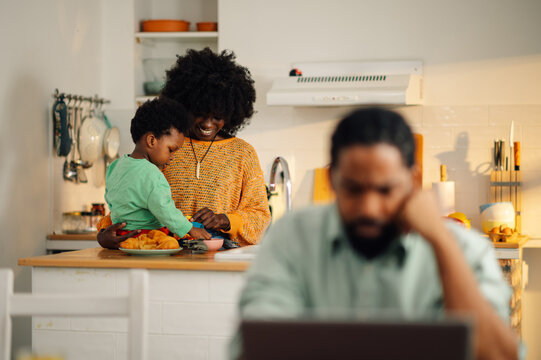 Selective focus on an african american woman with her son in the kitchen with her busy husband looking at the laptop in a blurry foreground.