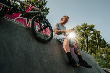 Bottom view of a middle-aged tattooed urban man typing messages on the phone in a skate park.