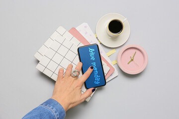 Female hand, mobile phone with text BLUE MONDAY on screen, cup of coffee and office supplies on grey background
