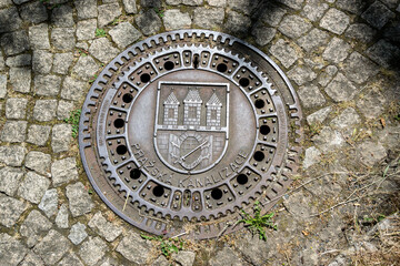 Iron canal lid in Prague, Czech Republic with the coat of arms and coat of arms of the capital. A channel on a cobblestone sidewalk.