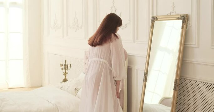 In the serene ambiance of a bedroom, a plus-size Caucasian woman, wearing a robe and lingerie, stands beside the mirror, gazing at her reflection with a joyful expression.