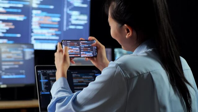 Close Up Back View Of Asian Female Programmer Looking At Database On Smartphone While Writing Code By A Laptop Using Multiple Monitors Showing Database On Desktops In The Office
