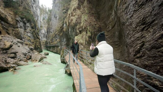 A blonde girl in hat and warm white vest takes a picture on her phone of a lady in jacket walking towards her on the wooden bridge, embraced by the majestic mountains of Aare Gorge, Switzerland.