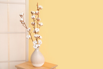 Vase of cotton sprigs on wooden stool with dressing screen near yellow wall