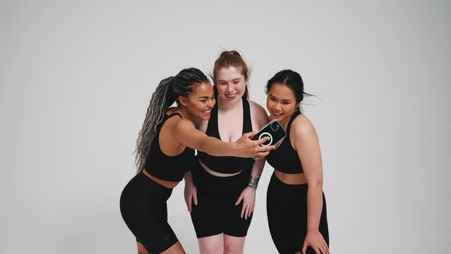 Three smiling multinational girls in black tracksuits take selfie photo on mobile against gray background in studio