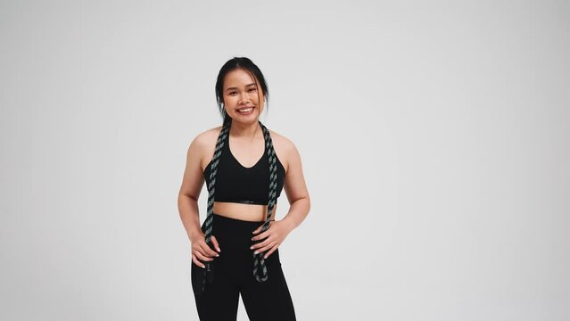 Positive asian woman holding skipping rope and greeting at camera in black tracksuit against gray background in studio 
