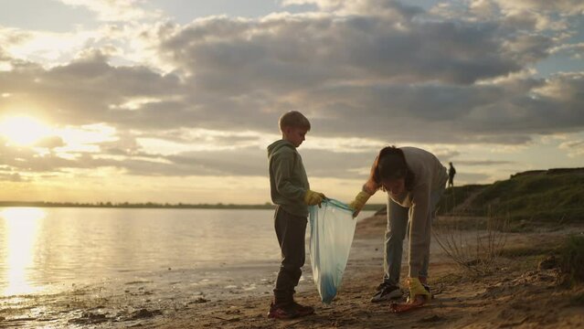 Mother And Son Cleaning Shore Of River, Picking Up Plastic Waste And Putting Into Bag For Recycling