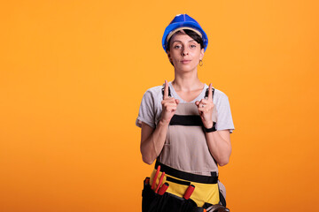 Portrait of woman builder pointing upwards with index fingers, showing above and indicating direction up. Posing in studio over yellow background and wearing hardhat with overalls.