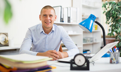 Portrait of smiling successful businessman sitting at workplace in office, posing during working...