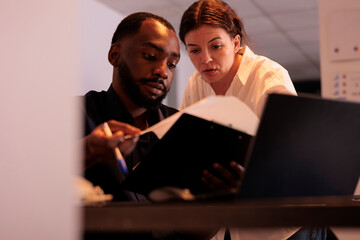 Project manager helping coworker checking report statistics, comparing data on laptop in office at night. Two employees checking marketing research results together in coworking space