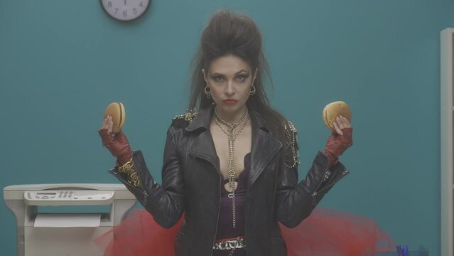 A woman stands in the office holding a hamburger in her arms. The woman takes turns greedily taking bites from both burgers, stuffing her mouth full. Advertising. Slow motion. HDR BT2020 HLG Material