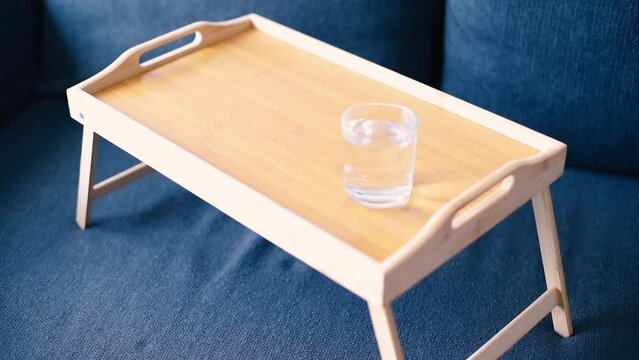 Una bandeja de madera sobre un sofá, en ella una mujer coloca un vaso con agua, un bol de noodles y un tenedor. Concepto de comer desde el sofá viendo la televisión gracias a una bandeja con patas.
