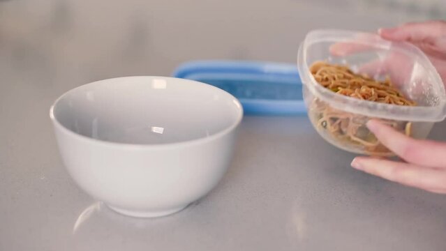 Abriendo un tupperware y vertiendo fideos en un bol sobre la encimera de una cocina. Concepto de conservar comida preparada en un envase de pl&aacute;stico.