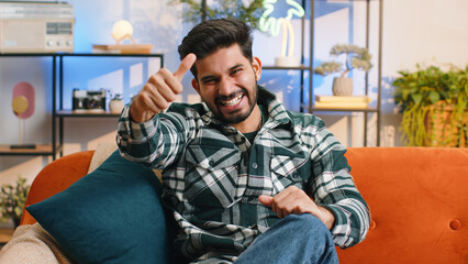 Happy indian man shouting, celebrating success, winning, goal achievement good news raising thumb up in like gesture, lottery luck at modern home apartment. Hindu guy in living room sitting on couch