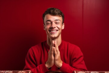 Medium shot portrait photography of a grinning boy in his 20s putting hands together as if praying against a ruby red background. With generative AI technology
