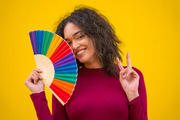Portrait of a latin woman smiling with a rainbow lgbt fan on a yellow background, making the victory symbol