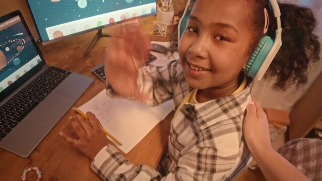 Handheld POV of mother recording vlog of her African American preteen daughter drawing at desk in her cozy bedroom in evening, smiling and waving at camera