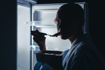 A woman with a short haircut eats near the refrigerator at night. Night appetite, diet concept. A...