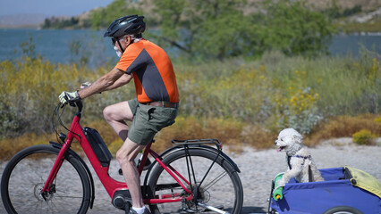 Obraz premium Elderly senior man biking on an e-bike on a trail pulling a trailer with a dog in it.