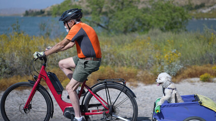 Elderly senior man biking on an e-bike on a trail pulling a trailer with a dog in it.