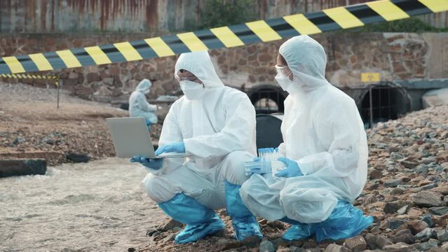 Ecologist sampling water from the river with test tube and save data to laptop computer, Biologist in a protective suit and mask collects sample of waste water from industrial, problem environment