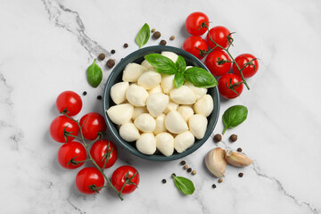 Delicious mozzarella balls in bowl, tomatoes and basil leaves on white marble table, flat lay