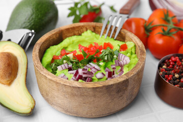 Bowl of delicious guacamole with onion, tomatoes and ingredients on white tiled table, closeup