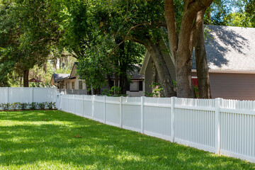 A stark white wood residential picket fence encloses a garden with tall green maple leaf trees on...
