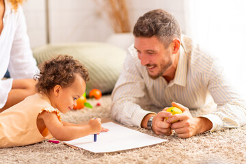 Obraz na płótnie Canvas Happy family parents and little daughter playing and coloring in a book together in living room at home. Father and mother teaching child drawing picture. Parenting and family relationship concept.