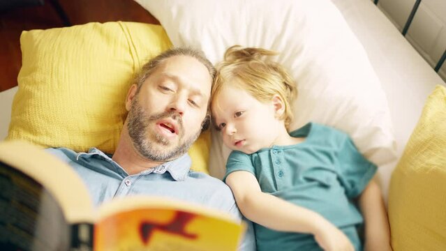 Caucasian father reading a book to little baby son on the bed in bedroom. Happy parent and child boy kid enjoy and fun indoors activity lifestyle together at home. Family love relationship concept.