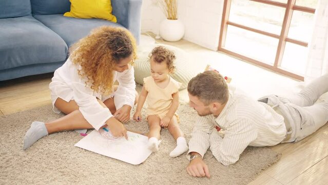 Happy family parents and little daughter playing and coloring in a book together in living room at home. Father and mother teaching child drawing picture. Parenting and family relationship concept.