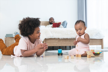 Obraz premium African family, little boy and cute toddler baby girl, brother and sister have fun playing model toys and wooden blocks together on floor while single dad lying on bed, reading book, looking care kids