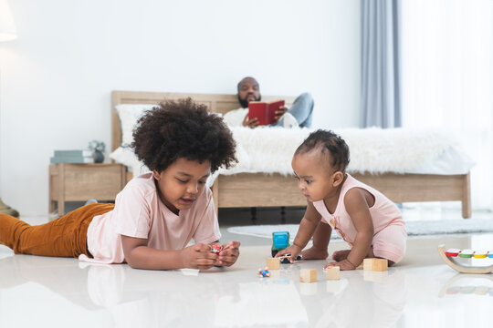 African family, little boy and cute toddler baby girl, brother and sister have fun playing model toys and wooden blocks together on floor while single dad lying on bed, reading book, looking care kids