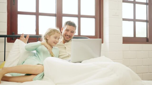 Caucasian father and little daughter resting on the bed in bedroom with using laptop computer play learning games or watch movie together. Happy family enjoy weekend activity with technology at home.