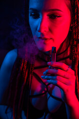 Caucasian girl with dreadlocks smokes a vape in red blue light. 