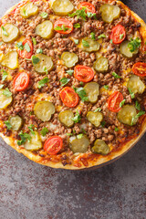 Delicious hot hamburger pizza with ground beef, tomatoes, pickles and cheese close-up on a wooden board on the table. Vertical top view from above