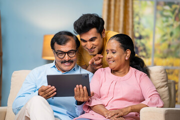 Indian senior parents with adult son watching digital tablet by talking together at home - concept of communication, parenthood and interaction.