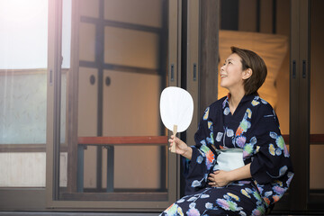 A middle-aged woman with an uchiwa, wearing a yukata, looking up and relaxing on the porch of a...