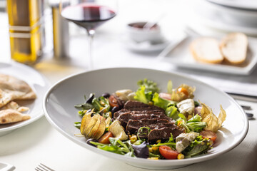 Beef steak sliced on top of mixed salad with greens, physalis, cherry tomatoes served with wine