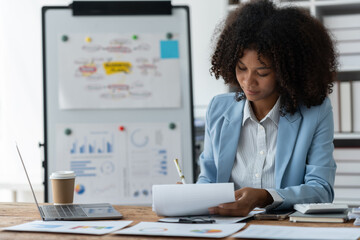 Project revenue and profit accounting African american businesswoman working with laptop financial charts and graphs checking and reading data details in financial paper documents at desk.
