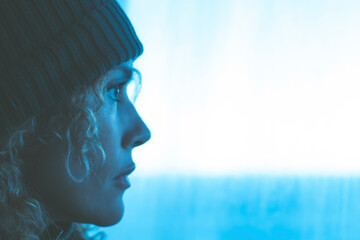 Side portrait of woman with knit hat looking outside the window in blue light. Travel adventure concept people lifestyle. Adult female dreaming and thinking with white copyspace background. Thoughtful