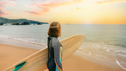 Unrecognizable young surfer woman with wetsuit carrying surfboard looking to the sea at the beach