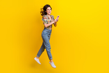 Fototapeta na wymiar Full body cadre of young girl jumping wear flowers print blouse jump use phone internet connection fast speed isolated on yellow background