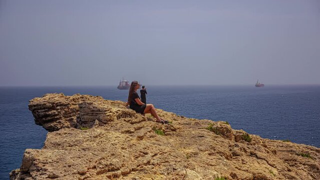 Timelapse shot of a couple sitting on stony cliff along sea shore with fishing boats in the background on a sunny day.