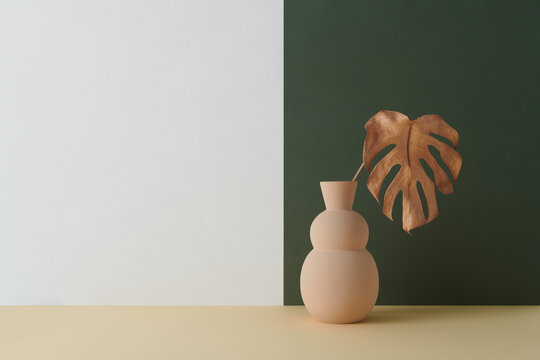 Horizontal shot of contemporary color-blocking still life composition of beige vase with dried Monstera leaf against white and dark green wall background, copy space